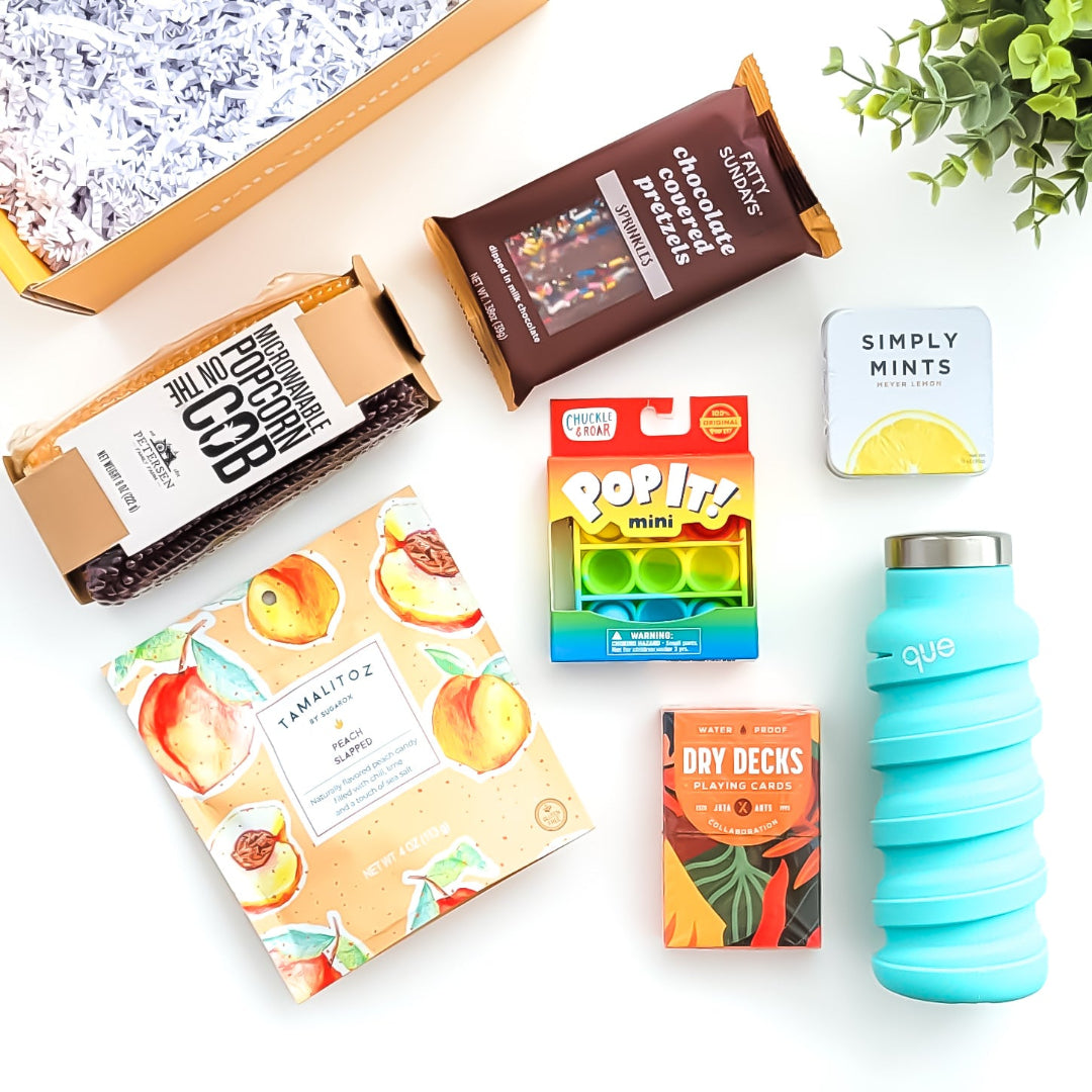 Let’s Play Gift Box featuring a fun game night collection, including microwaveable popcorn on the cob, peach candies, waterproof playing cards, lemon mints, a collapsible water bottle, a mini pop-it, and chocolate-covered pretzels—perfect for friends, family, and game lovers.